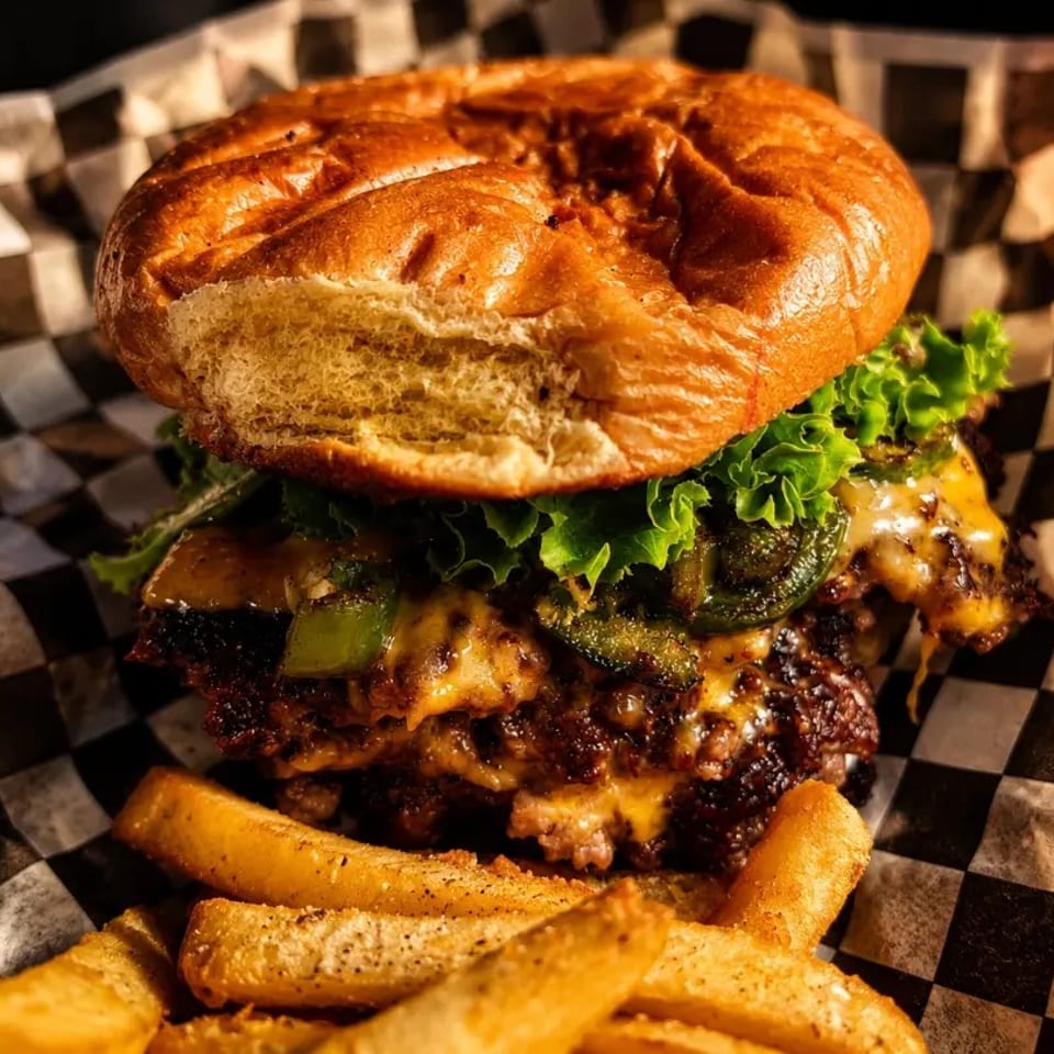 Cheeseburger with lettuce, jalapeños, and melted cheese, served with fries on checkered paper.