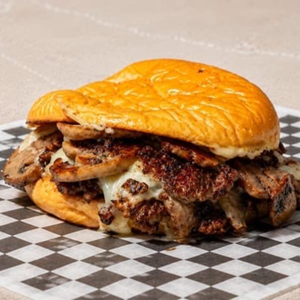 A hearty sandwich with meat, mushrooms, and melted cheese on a bun, resting on checkered paper.